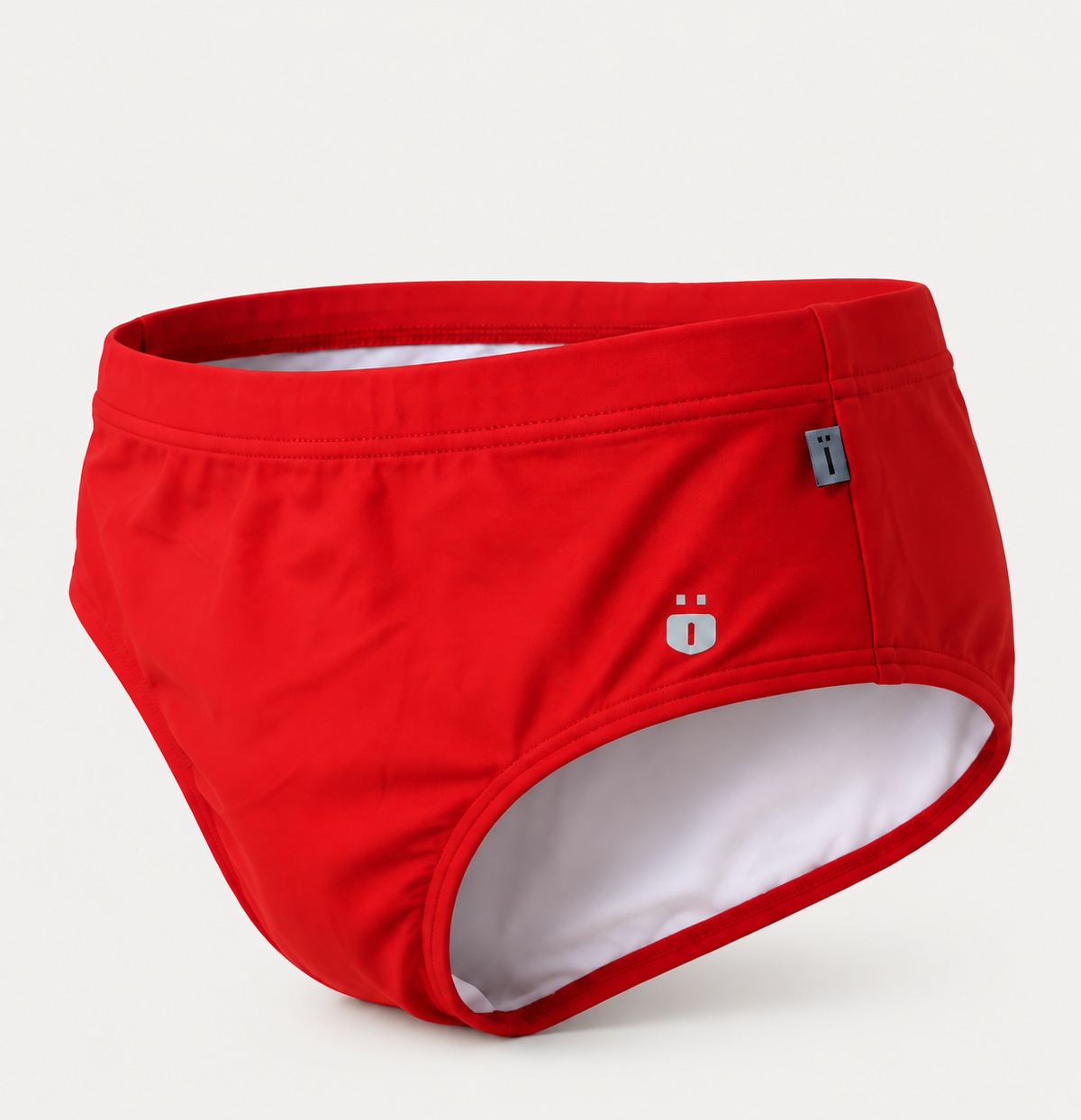 Red swim briefs with a white interior lining, featuring a small white logo and a gray brand tag on the side, set against a light gray background.